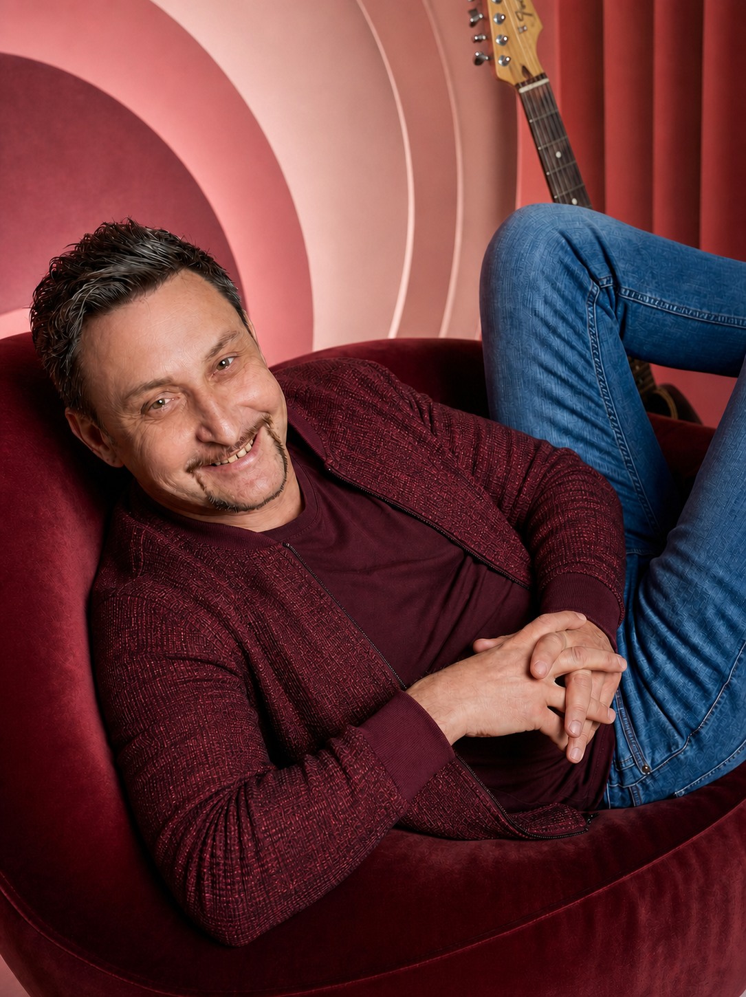 Denny Schönemann im weinroten Bühnenoutfit mit Gitarre vor roséfarbenem Studiohintergrund